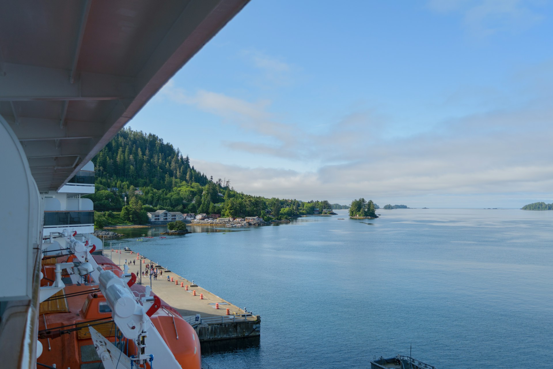 View from a cruise ship overlooking a calm bay with a small harbor, surrounded by green hills and trees under a blue sky.