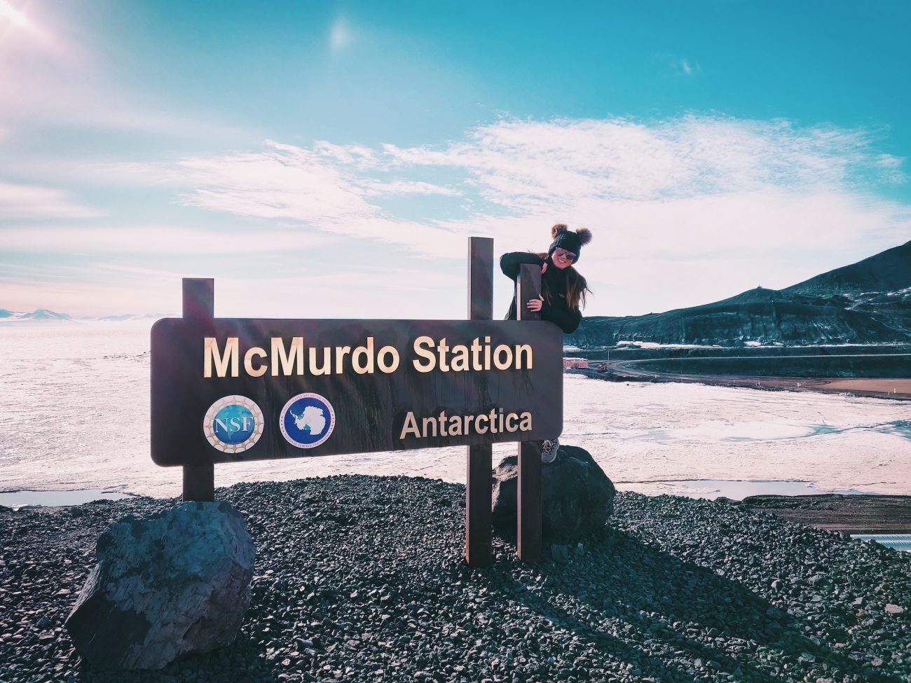 A girl stands next to the McMurdo Station sign in Antarctica-- another job that travels (no experience necessary)