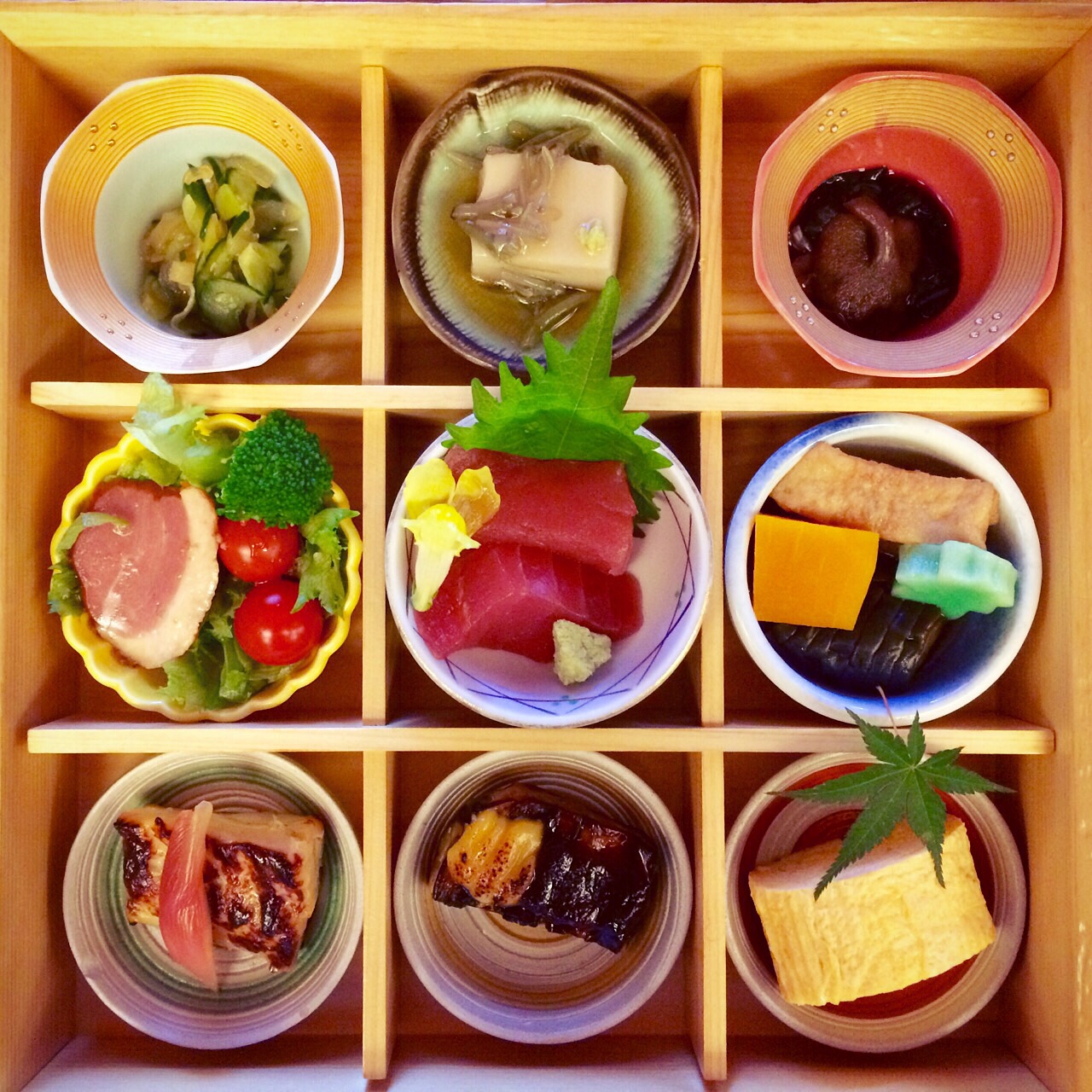 Japanese food in a wooden box split into 9 sections, each with a small dish 