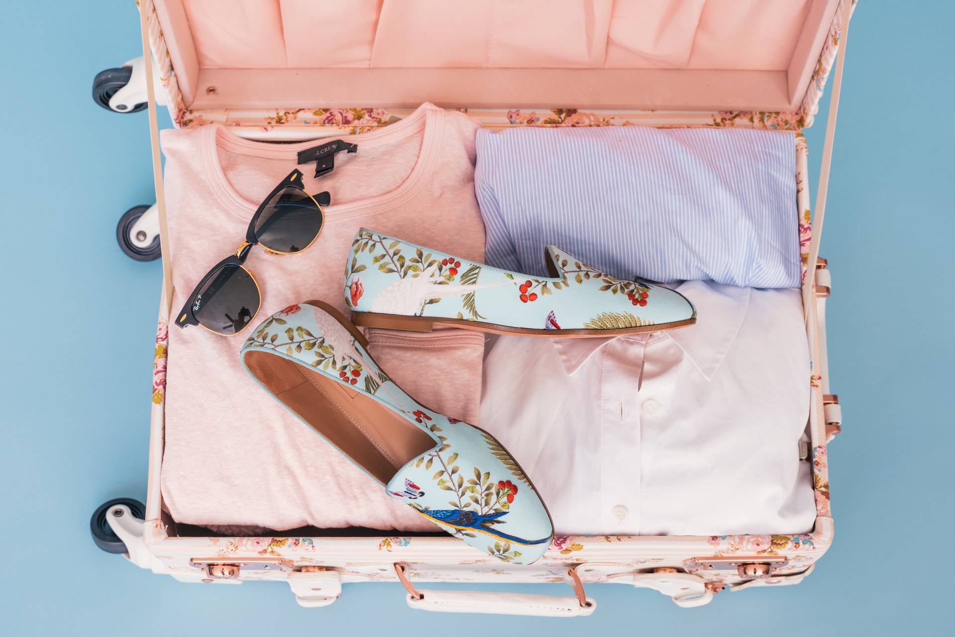 opened pink suitcase with pastel colored shirts, light blue floral slip on shoes, and sunglasses