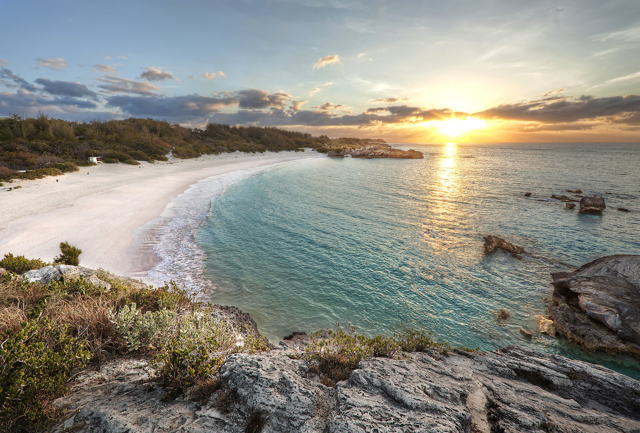 Sunrise at Horseshoe Bay in Bermuda, rocky cliffs in the foreground, turquoise water to the right, white sand beach to the left, and sunset in the far upper corner