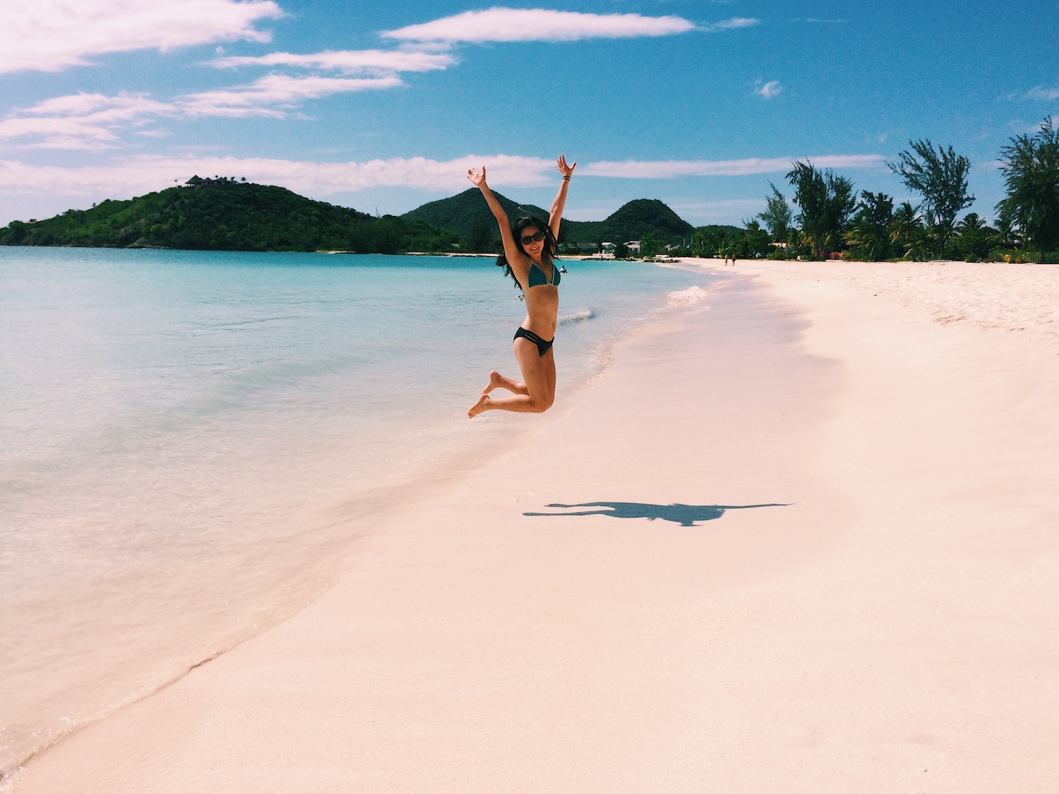 Jumping for joy on Jolly Beach in Antigua; background shows soft, white sand with electric blue waters and Caribbean mountains