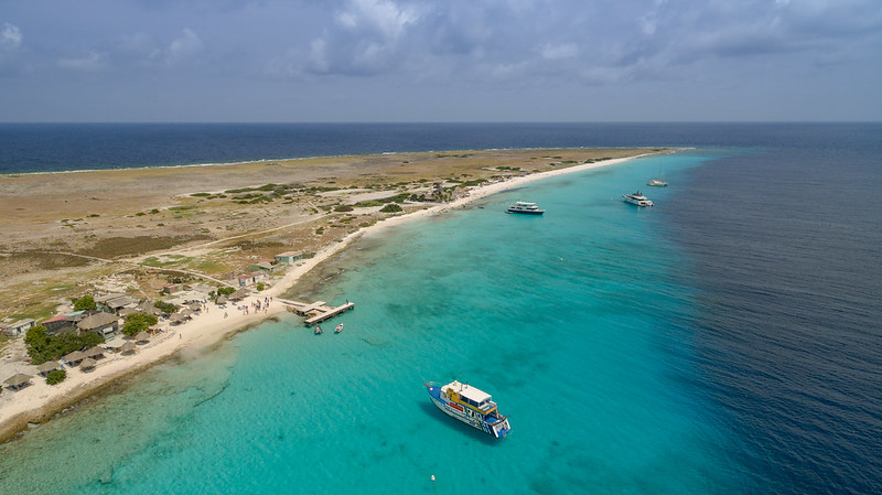 An aerial view of Klein Curacao, a 1.7 sq km uninhabited island with deep blue navy waters farthest from the island, turquoise water close to the island with a few small boats, and the island's sandy shore to the left of the photo 