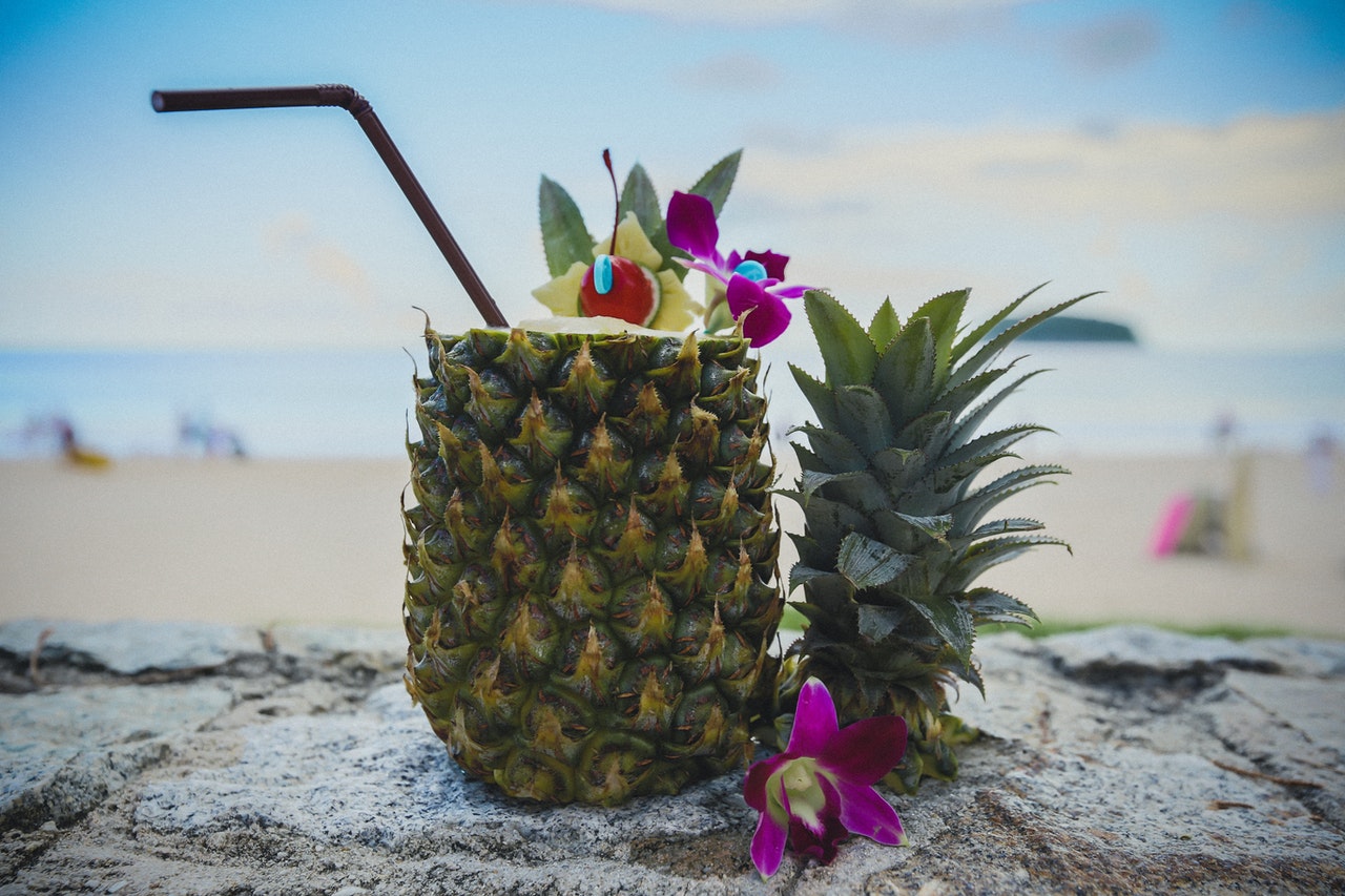 cocktail in cut open pineapple with floral garnish and a straw by the beach