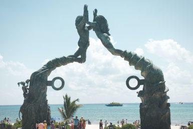 two large mermaid statues joining hands to form a gateway in front of the ocean; tourists walking in front of it
