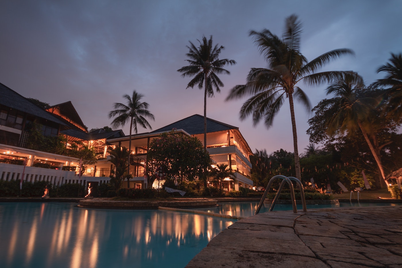 Night time view of exterior of resort with swimming pool, night lighting, and palm trees