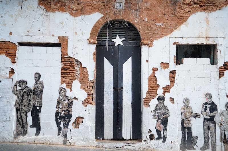 Mural of black and white puerto rican flag on door of abandoned building next to paintings of people