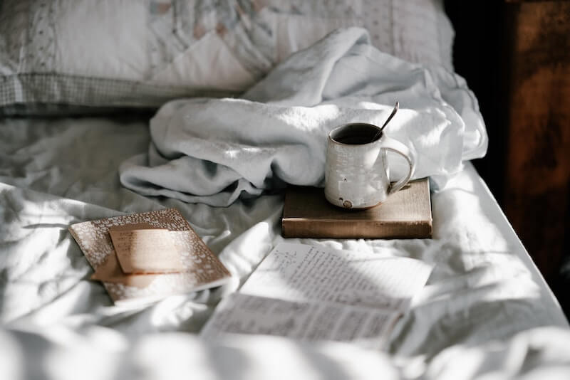 cozy bed with white sheets and blankets, a couple books, and mug with coffee