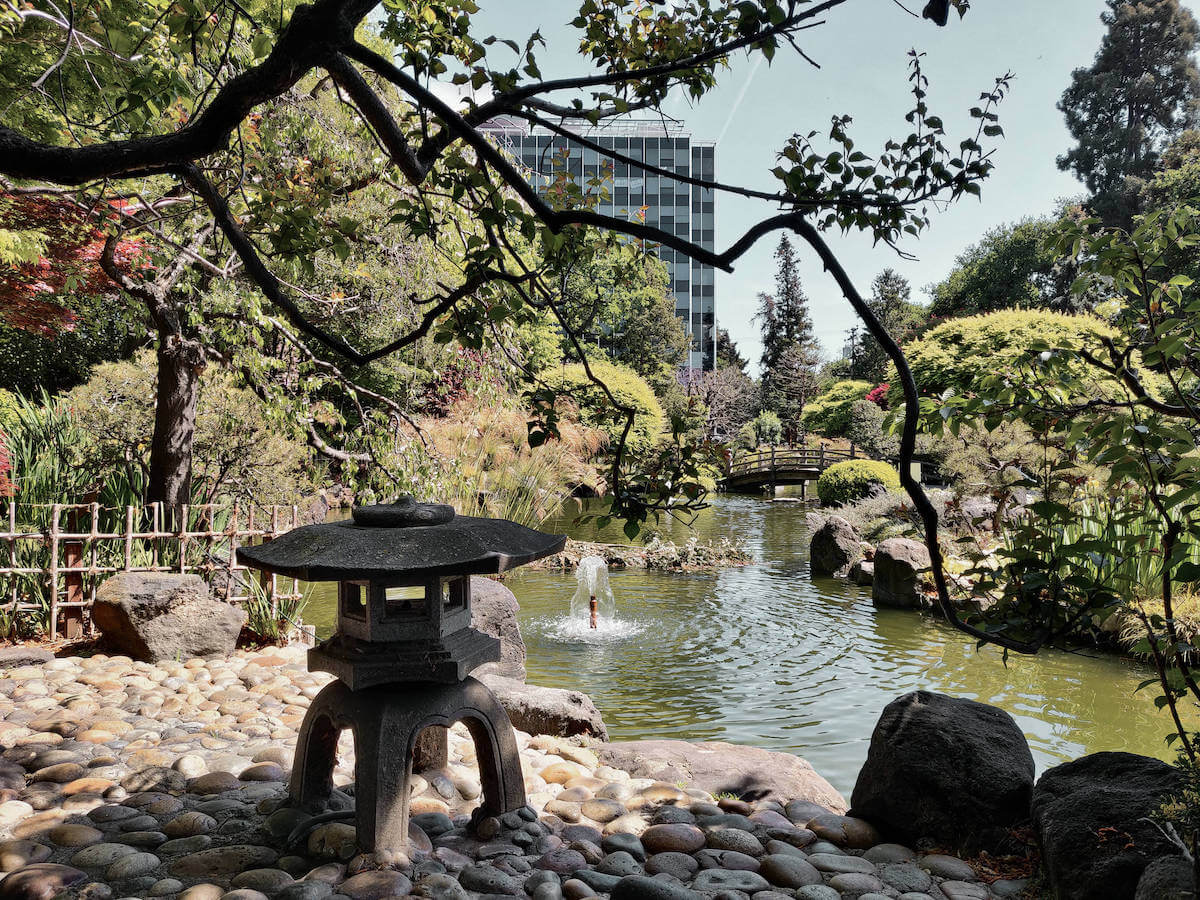 Japanese stone lantern in front of a koi pond and bridge with greenery around