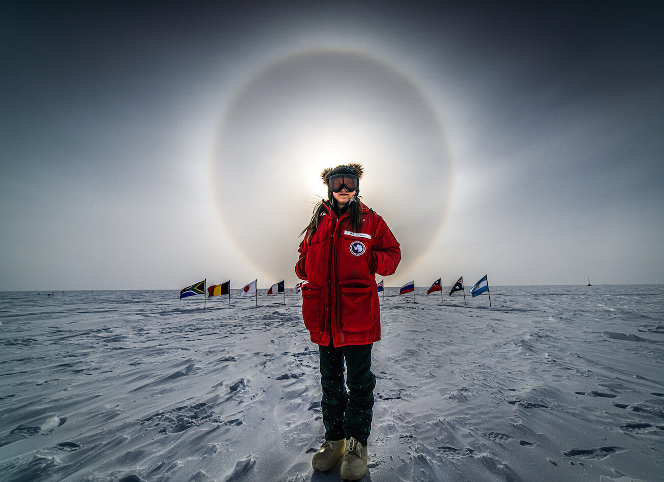 A woman stands in front of flags at the Ceremonial South Pole under a sun dog halo. She wears a Big Reg parka, black pants, white boots, tinted goggles, and a beanie.