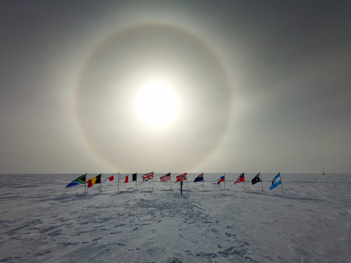 The Ceremonial South Pole with a sun dog overhead. 12 flags surround the pole marker from the following nations: South Africa, Belgium, Japan, France, Great Britain, United States, Norway, New Zealand, Russia, Chile, Australia, and Argentina