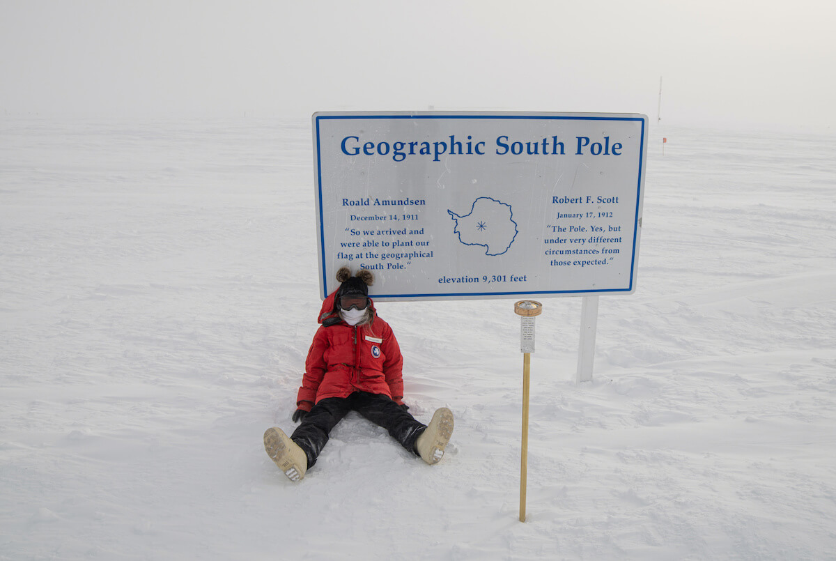 A girl wearing a Big Red USAP-issued parka, a black beanie with two brown pompoms, black pants, and white boots sits at the Geographic South Pole Sign. The 2021 pole marker is gold with names etched into a silver vertical plaque.