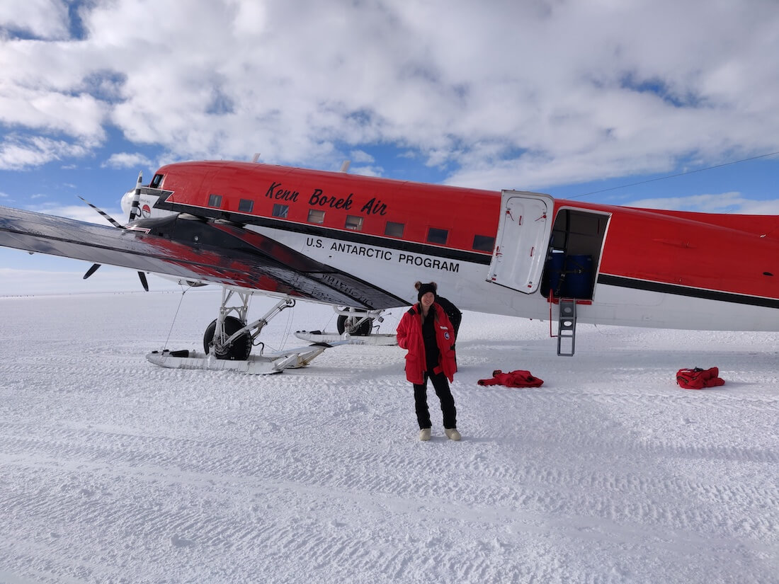 Kenn Borek Air modified DC-3 painted red and white with the U.S. Antarctic Program printed on the side. Michelle stands in front of the plane in her Big Red parka, black pants, white bunny boots, and beanie with pompoms.