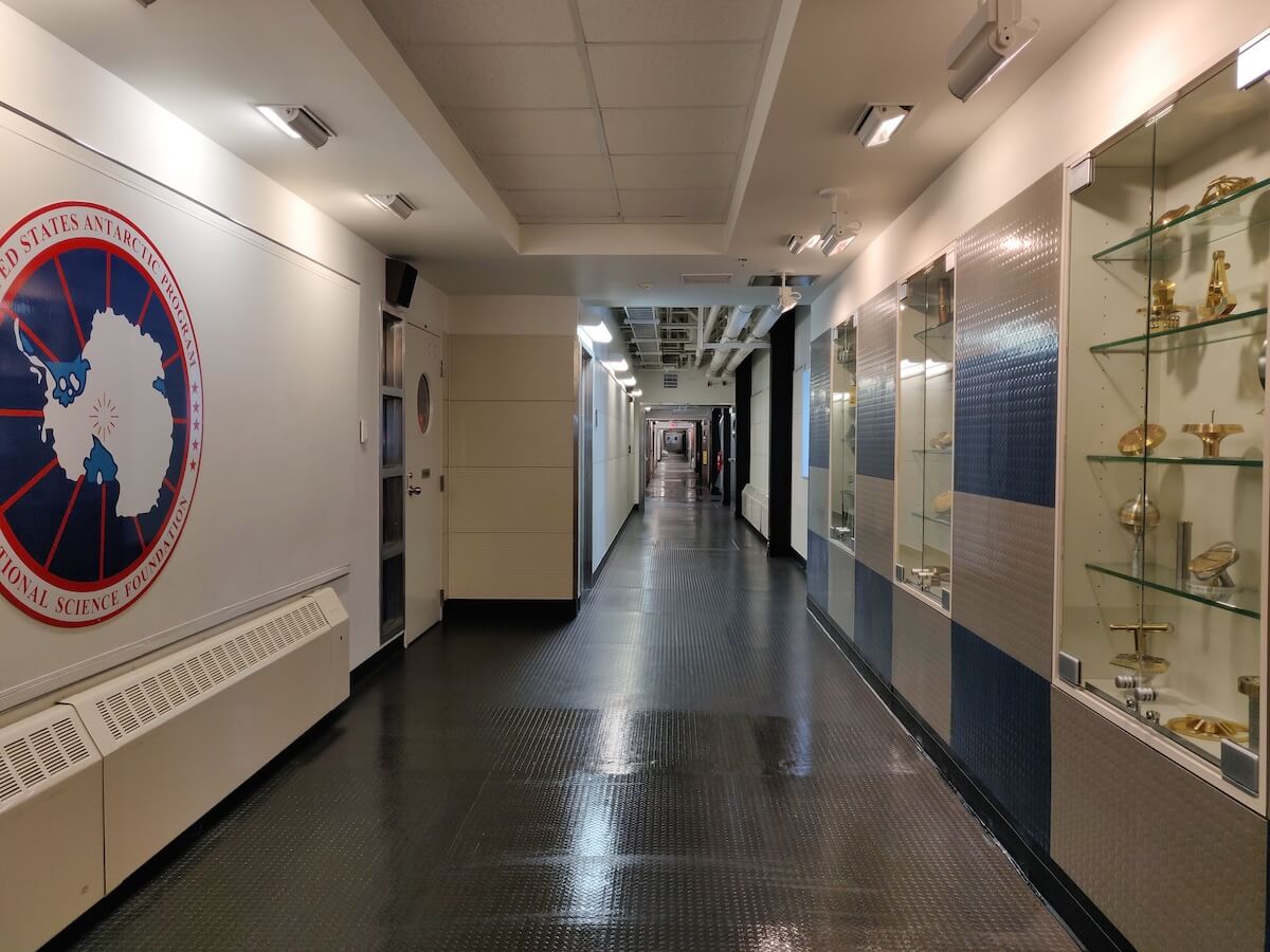 A long hallway with the USAP logo on the left and former South Pole markers encased on the right.