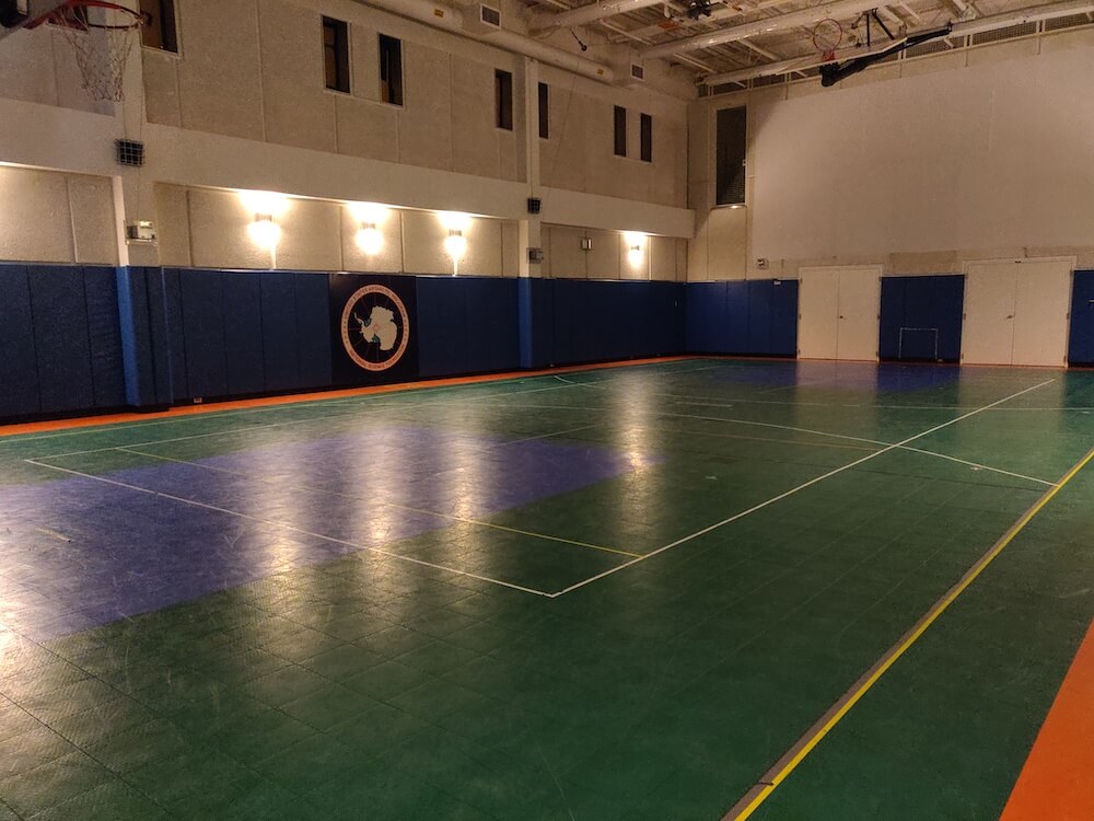 The South Pole gymnasium: green linolium floor with a basketball court marked with tape. The USAP logo is on the far blue wall