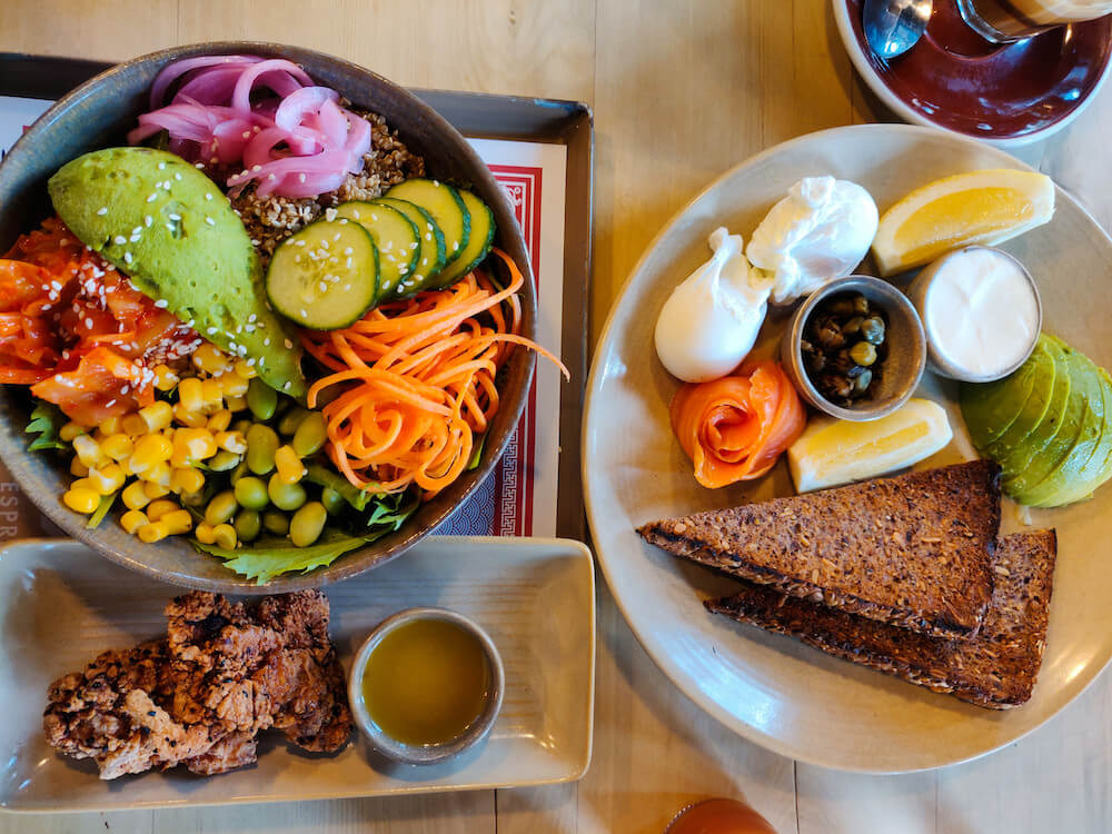 Two brunch plates from C1 Espresso: a Buddha Bowl with quinoa, carrots, corn, edamame, cucumbers, avocado, pickled onions, and fried chicken karaage on the side; second plate-- multigrain toast with smoked salmon, two poached eggs, slices of avocado, and lemon