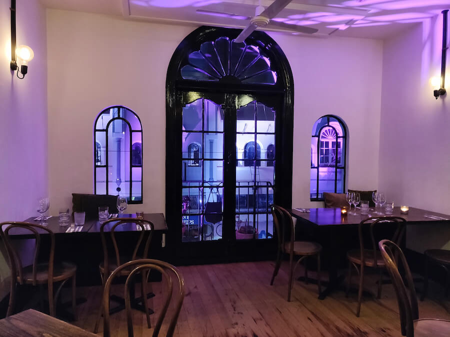 Cozy interior of Twenty Seven Steps Christchurch Restaurant: dark tables, dark hardwood floor, french doors leaving out to a purple-lit balcony at night