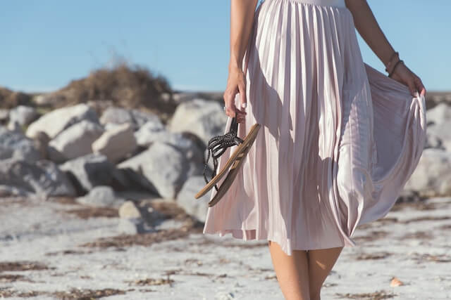 woman wearing a pleated, blush, midi-skirt walks on a beach holding brown beach sandals