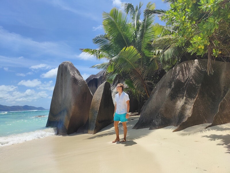 man stands on a beach in the seychelles in front of large granite rocks and palm trees. he wears a white linen short sleeve button down shirt and teal boardshorts