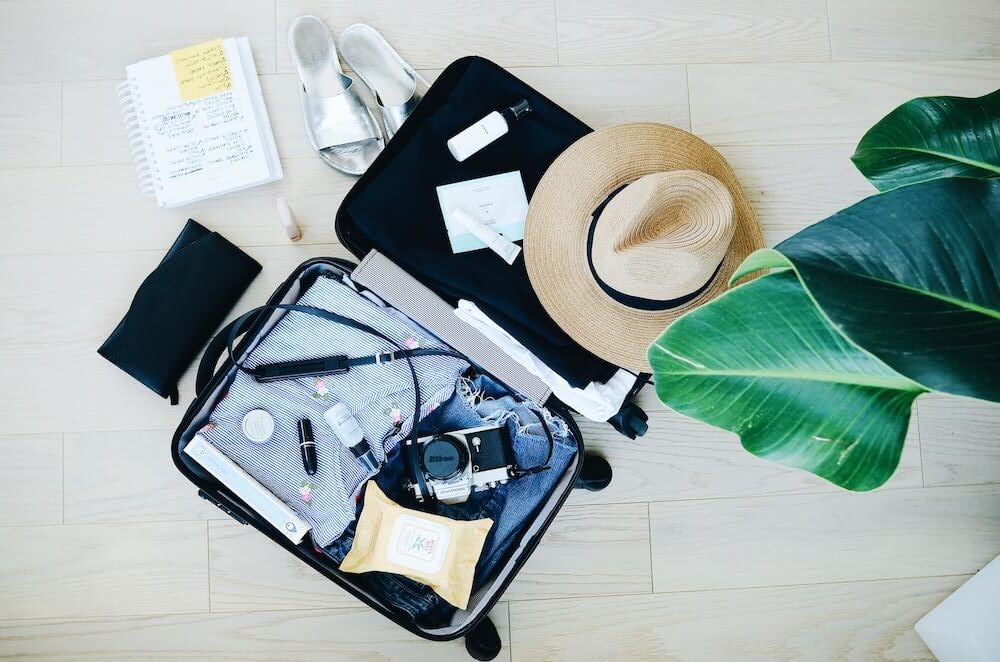 open suitcase with items shown in a flatlay: sun hat, cosmetics, blue top, denim shorts, camera, sandals, and a notebook