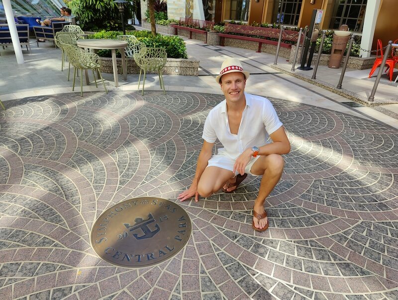 A man wears a straw fedora, white linen short-sleeve button down shirt, white linen shorts, a watch, and leather sandals. He squats next to the sign on the ground that says "Symphony of the Seas Central Park"