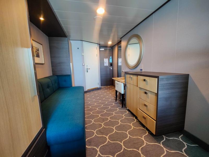 The interior of an ocean view balcony stateroom on Symphony of the Seas. Photo shows a wardrobe, sofa, dresser, and vanity area with chair and mirror