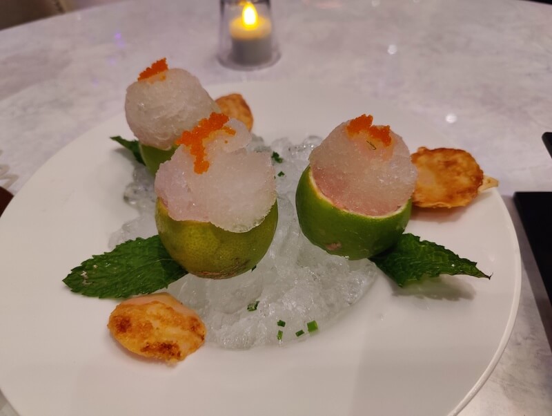 ice granules topped with small orange fish eggs sit in halved limes on a platter of ice with decorative leaves