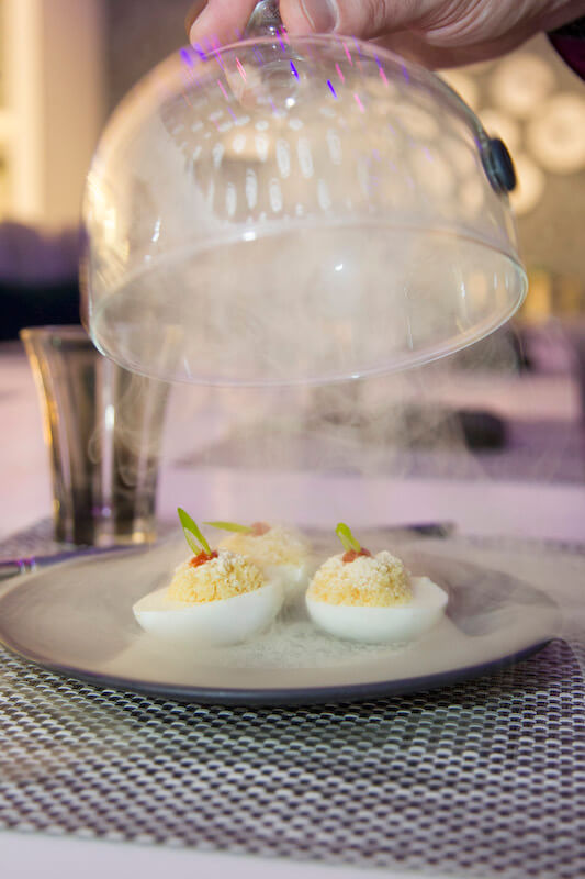 three half deviled eggs sit on a plate as a transparent dome is lifted. smoke is flowing out.