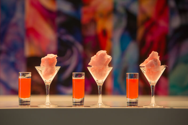 Three tall shot glasses filled with a red cocktail sit on a table between three martini glasses filled with cotton candy