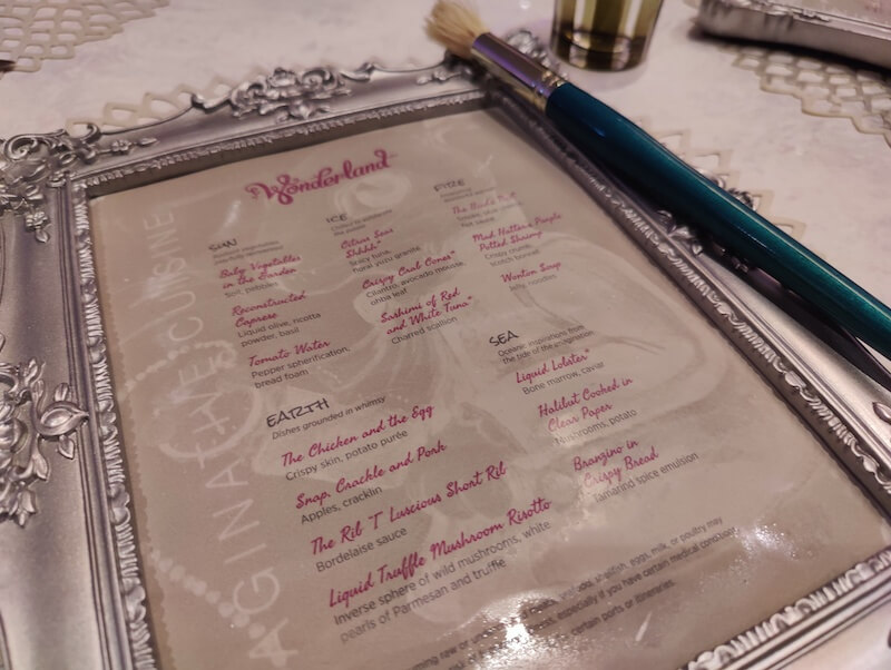 A framed Wonderland menu painted over with water to reveal the print. A large paintbrush sits to the right.