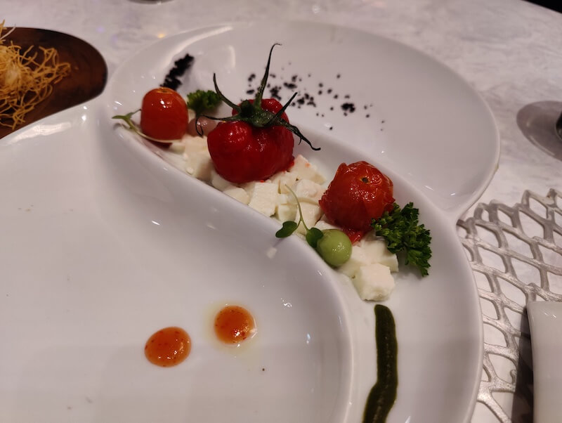 A white dish with a narrow valley in the middle holds small cubes of ricotta cheese and a reconstructed tomato that's been blended with herbs. Green garnishes sit to the side with a few drops of orange sauce.