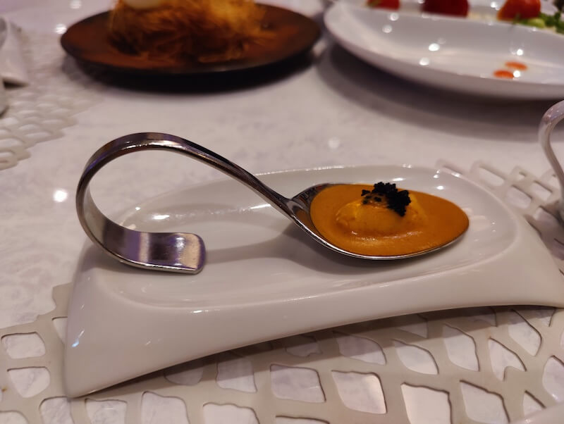 one bite serving of a piece of lobster and orange sauce topped with caviar sits in a whimsical curved silver serving spoon on a white serving dish
