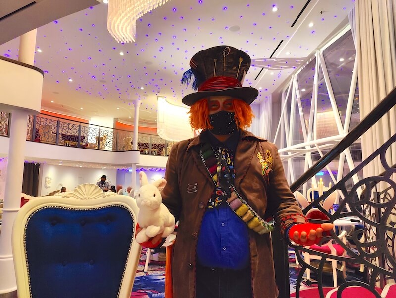 The Mad Hatter is dressed in a brown trench coat, blue shirt, and wears a colorful sash across his shoulder and under an arm. He has bright orange hair, wears a whimsical top hat, and red fingerless gloves. He also holds a plushie rabbit.