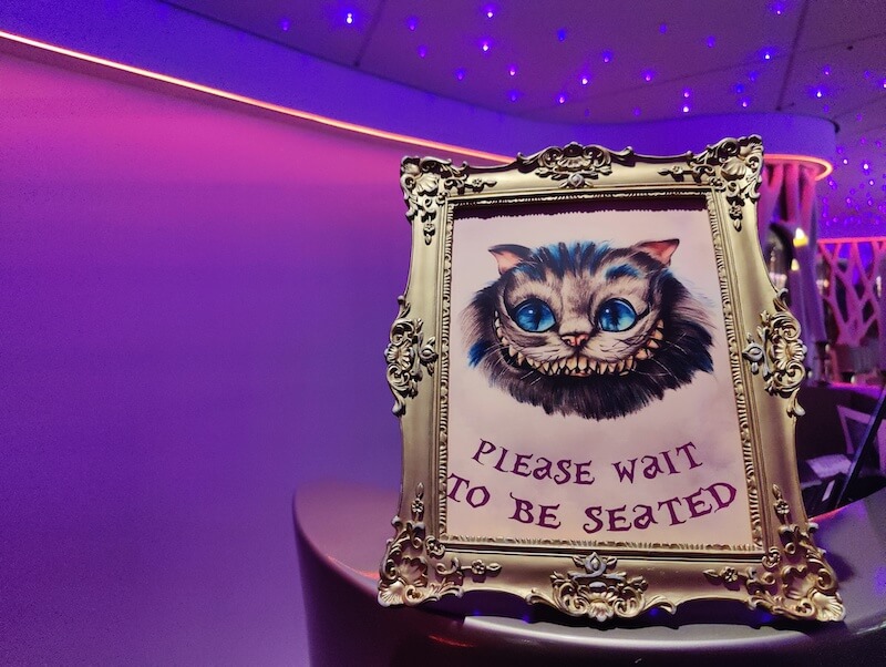 framed sign that says "please wait to be seated" with a picture of the cheshire cat