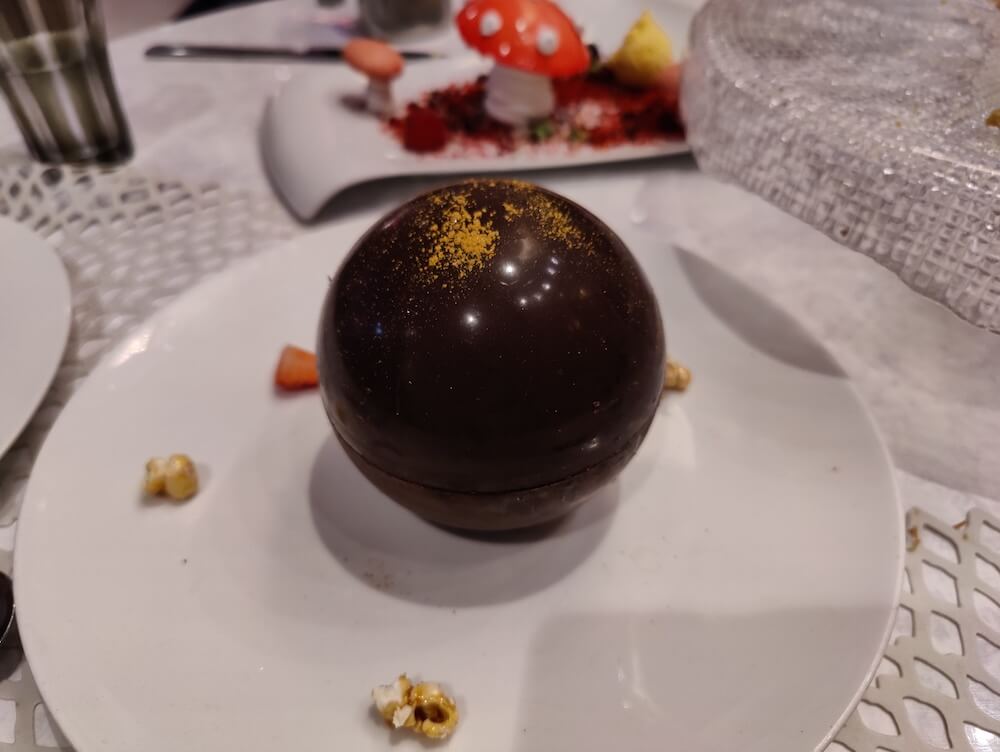 A chocolate orb dessert dusted with gold sits on a white plate