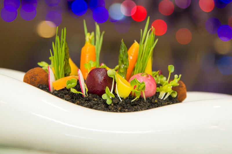 baby vegetables of radishes, greens, asparagus, and carrots appear to grow out of soil in a white dish that looks like a flower bed