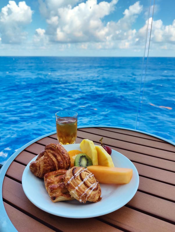 A plate of pastries and fresh assorted fruit slices with a glass of apple juice sit on a small table on a Symphony of the Seas balcony with a view of the ocean