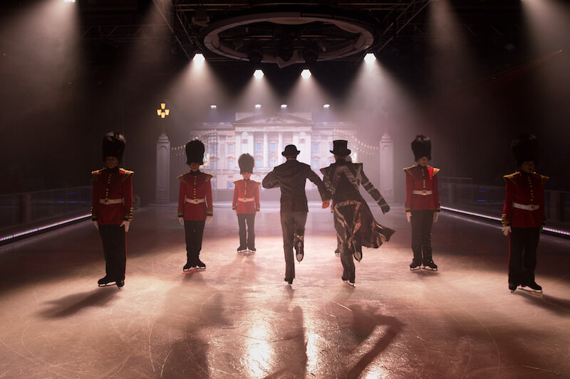 Ice skaters dressed like Buckingham Palace guards stand still as two men skate past on Symphony of the Seas' 1977 ice show