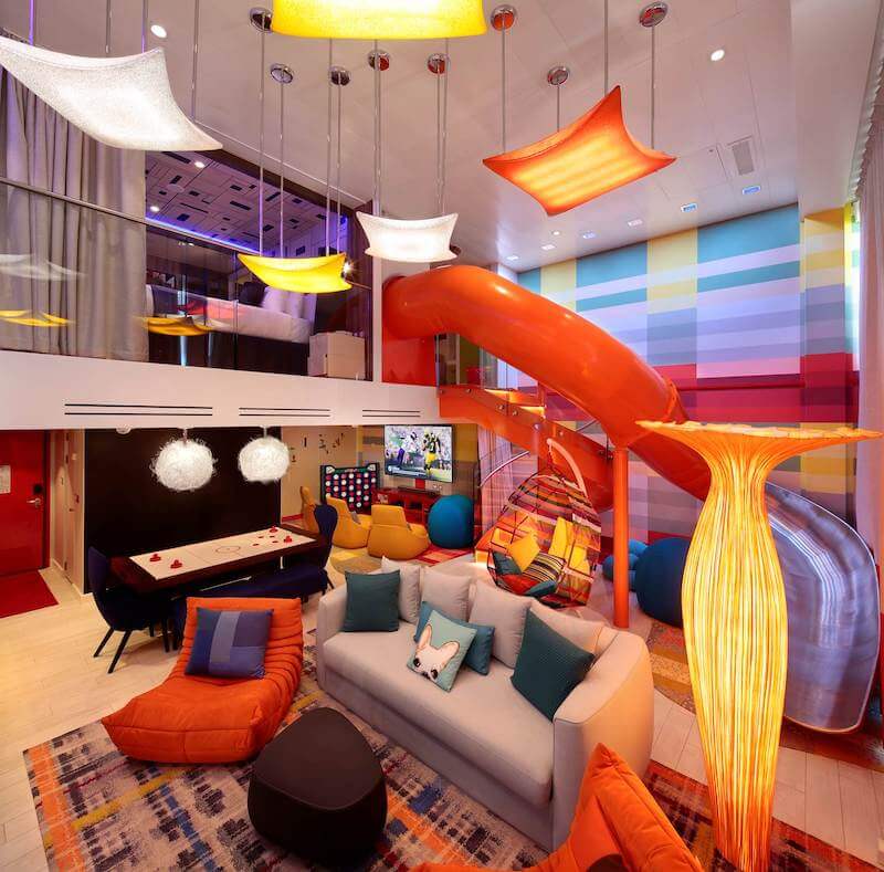 Interior of Symphony of the Seas' Family Suite. Includes a slide from the second to first floor, colorful decor and furniture, a TV theater, and air hockey table
