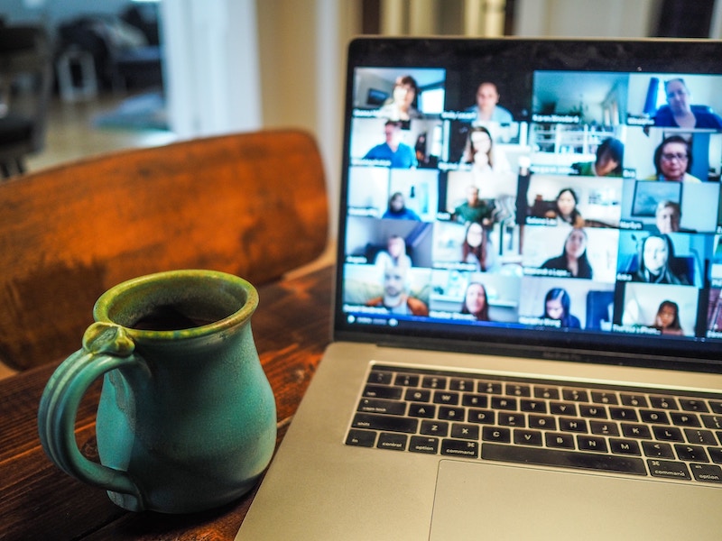 A laptop sits on a wooden table. The screen shows 25 people on a video call. A green mug is next to the laptop