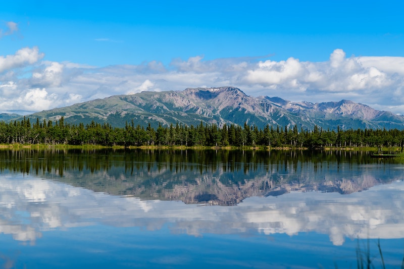 View of blue sky, clouds, mountains, and trees mirrored in the lake