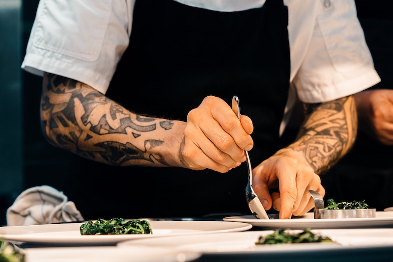 A chef with tattooed arms plates a dish with some greens