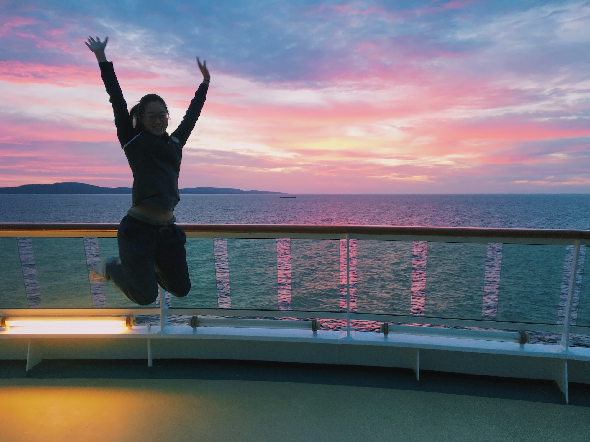 One of the best jobs that travel with no experience required: cruise ship worker. A female staffer jumps on the deck of a ship to pose during sunset