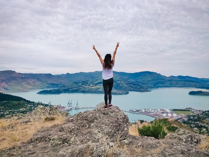 A girl stands on a rock overlooking a lake and mountains in New Zealand. Getting a working holiday visa in New Zealand is one of the best jobs that allow you to travel with no experience
