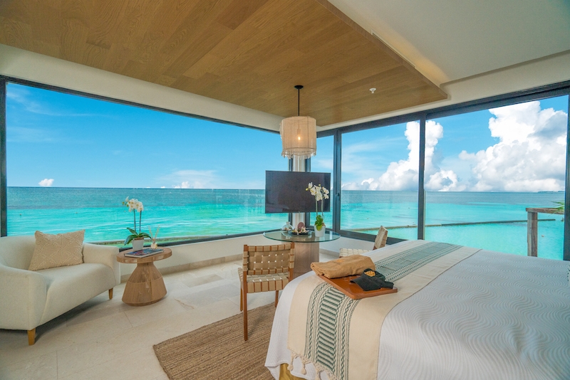 Inside a Palafitos Overwater Bungalows suite in Playa del Carmen, an adults-only all-inclusive resort. Photo features floor-to-ceiling glass windows with panoramic views of the water, a bed, sofa chair, and tv.
