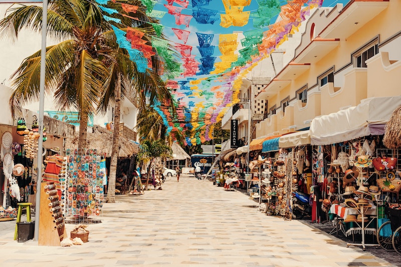 A street along Quinta Avenida in Playa del Carmen, one of the best things to do in the area. The street is covered with colorful banner cut outs. On both sides of the pedestrian street are shops with various displays such as hats.
