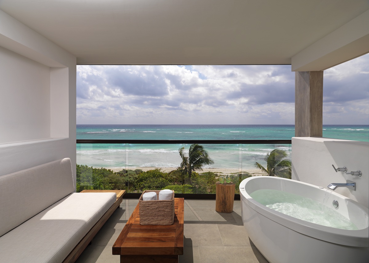 Photo of a private terrace at Unico 20°87° all inclusive Playa del Carmen adult only resort. Photo shows an outdoor private soaking tub, lounge bench, table with towels in a basket, and a glass balcony with a view of the ocean