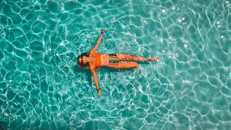 A woman in an orange bikini top and striped colorful bikini bottoms floats in a clear, blue swimming pool