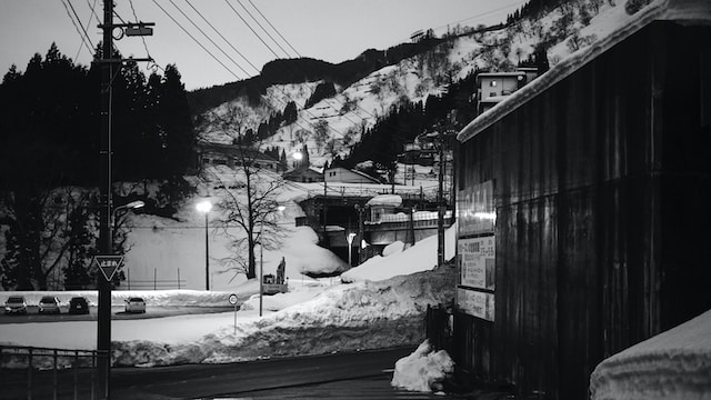 A black and white photo of Niigata, Japan: a parking lot with a few cars, telephone lines, and a building are surrounded by snow along a mountain
