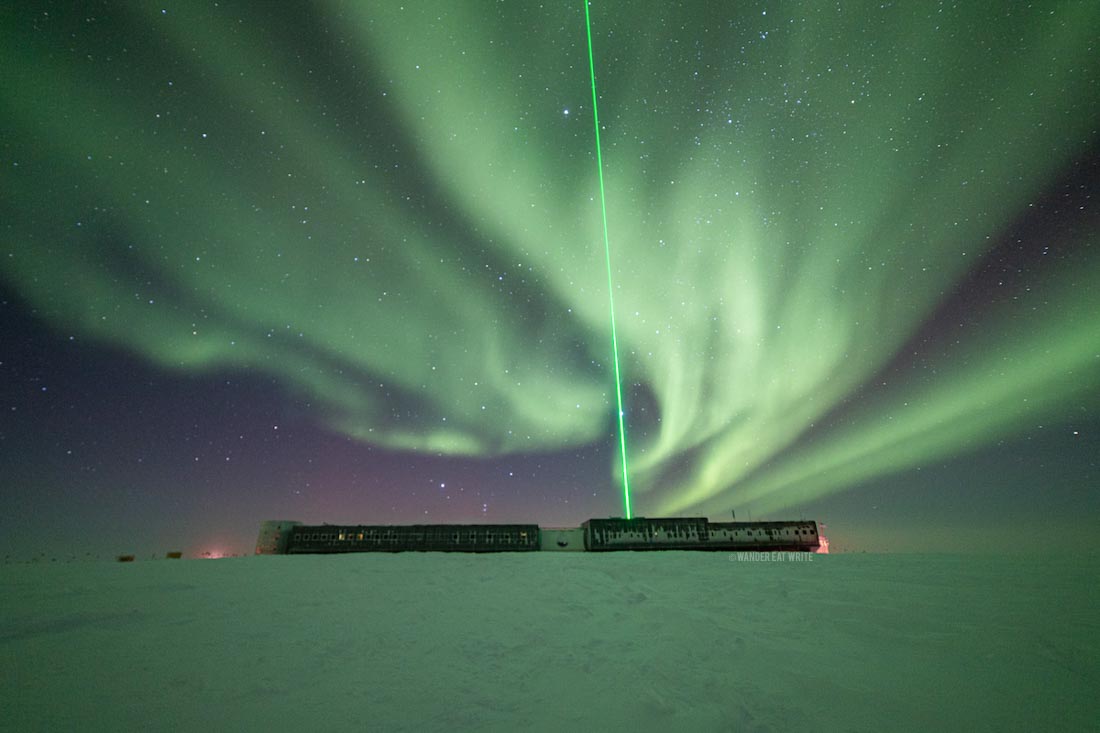 A green aurora filling the winter night sky above the Amundsen-Scott South Pole Station. The icy ground is lit up in green and a green laser beams up from the station